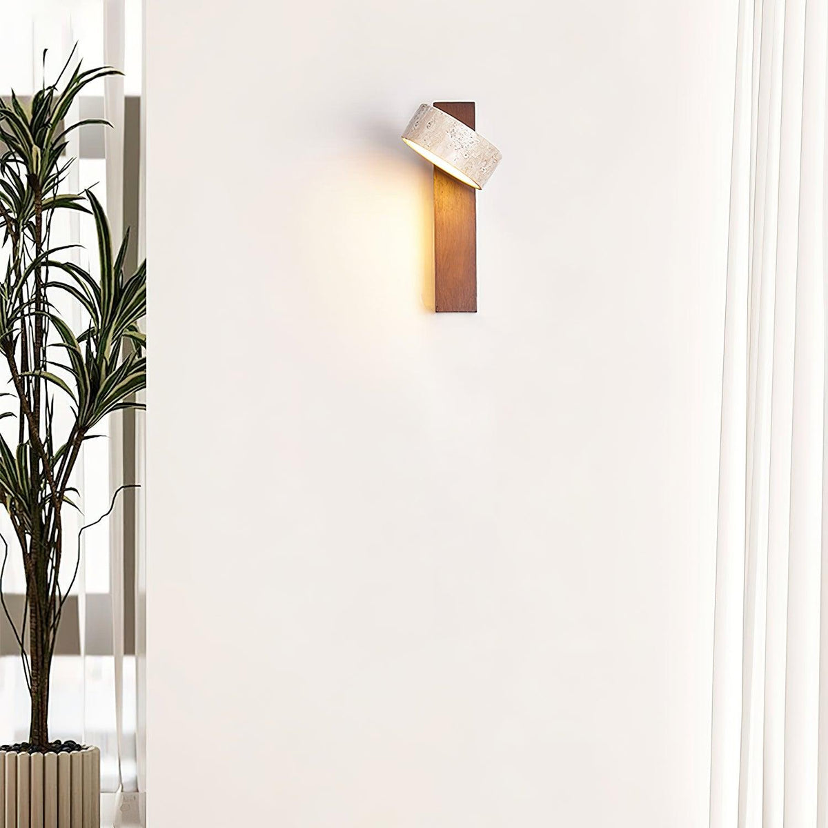 Nydo travertine wall light lit on a pale wall beside a tall indoor plant