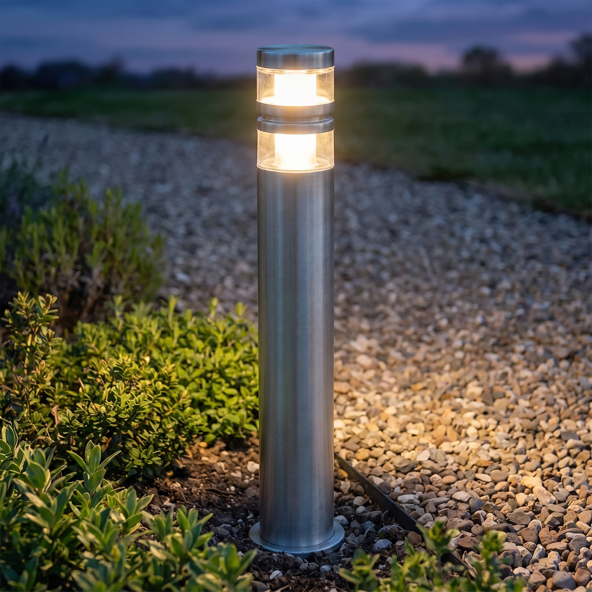 Vestro path post light glowing in a planted border beside a gravel garden path at dusk