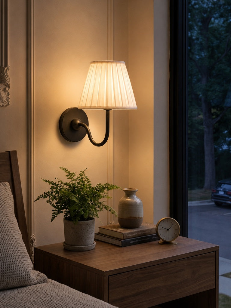 Nora wall light lit in the evening beside a bed, casting a warm pool on the wall