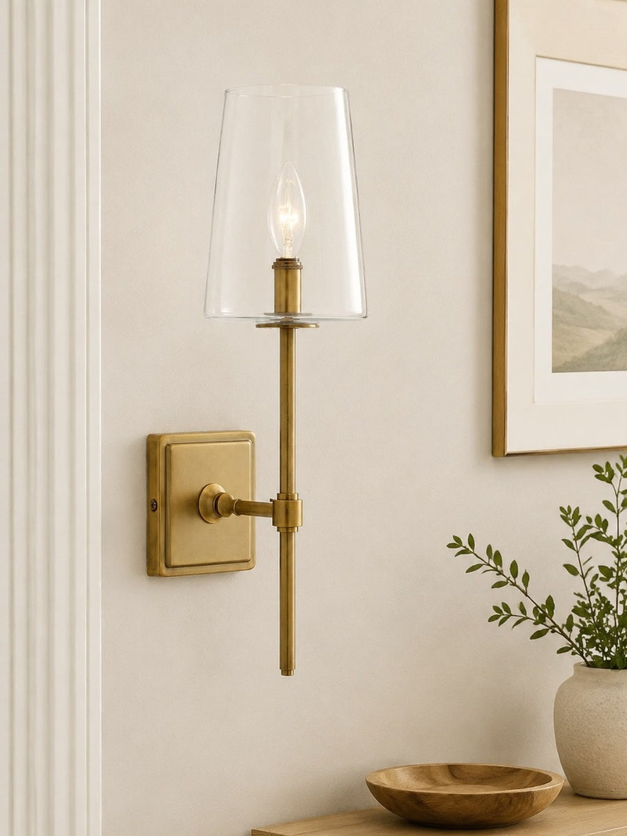 Torin Clear Glass Wall Sconce in tumbled brass mounted on a cream wall beside framed artwork