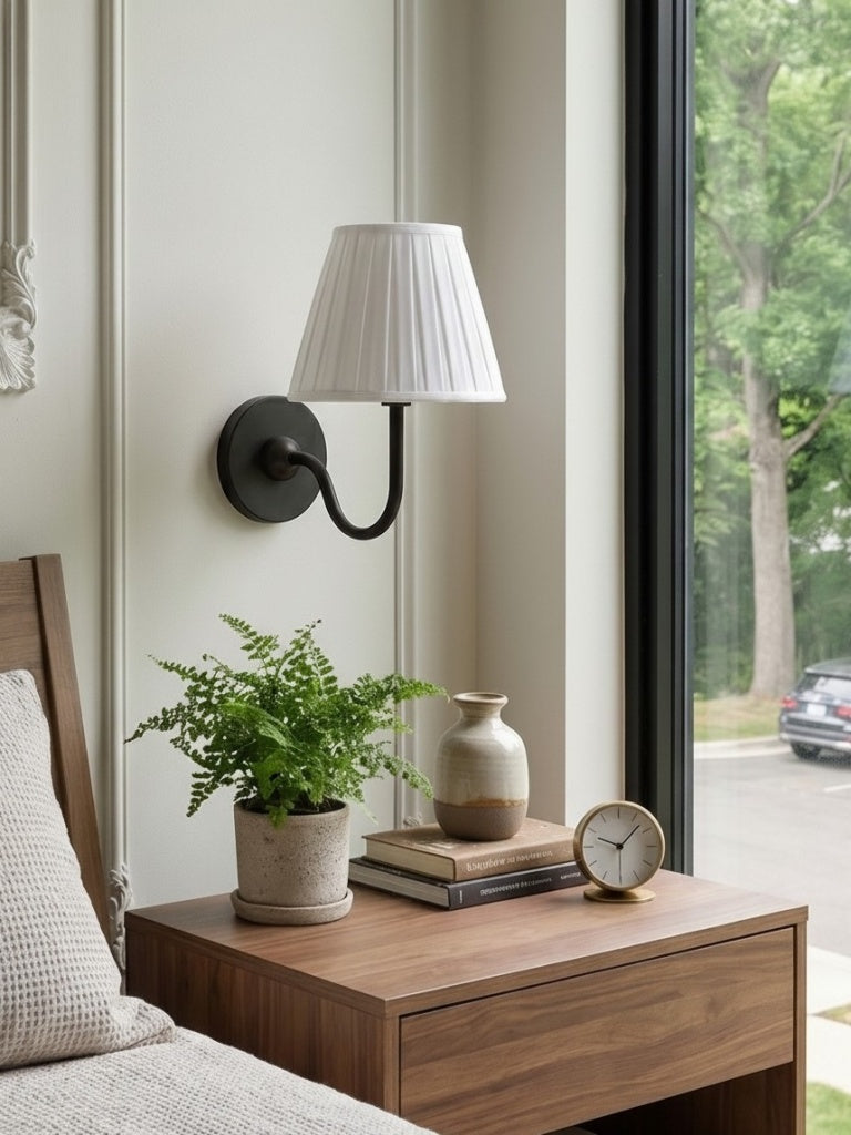 Nora matte black wall light with white pleated shade above a walnut bedside table in daylight