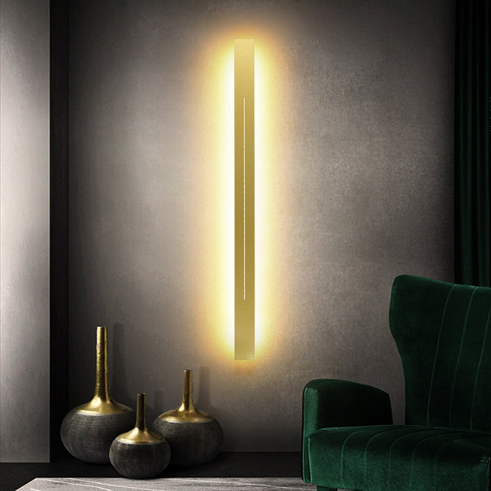Azino Slim LED Wall Light glowing warmly on a dark concrete wall beside a green velvet armchair