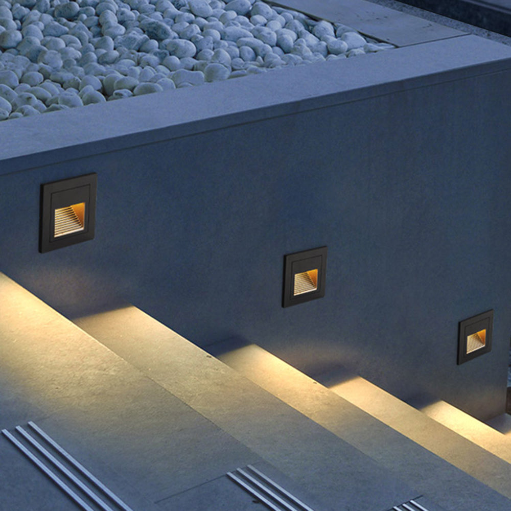 Three Steva square step lights recessed into a grey garden wall, washing warm light across concrete steps