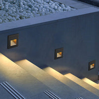 Three Steva square step lights recessed into a grey garden wall, washing warm light across concrete steps