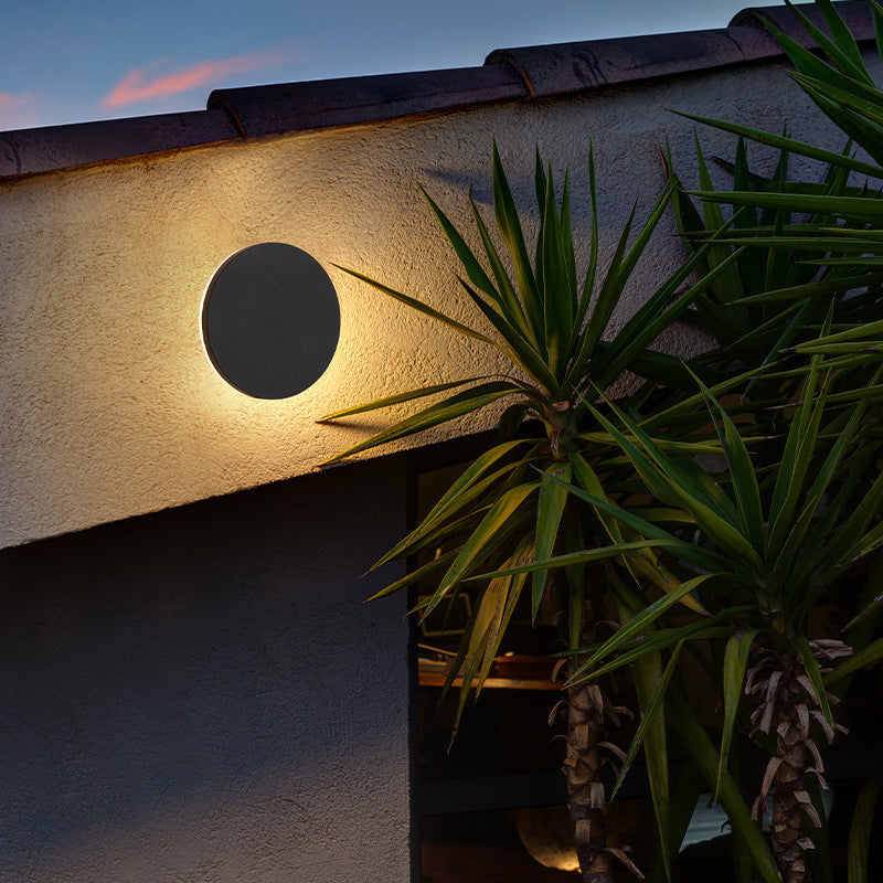 Klyno black round outdoor wall light lit at dusk on a rendered house wall beside a yucca plant