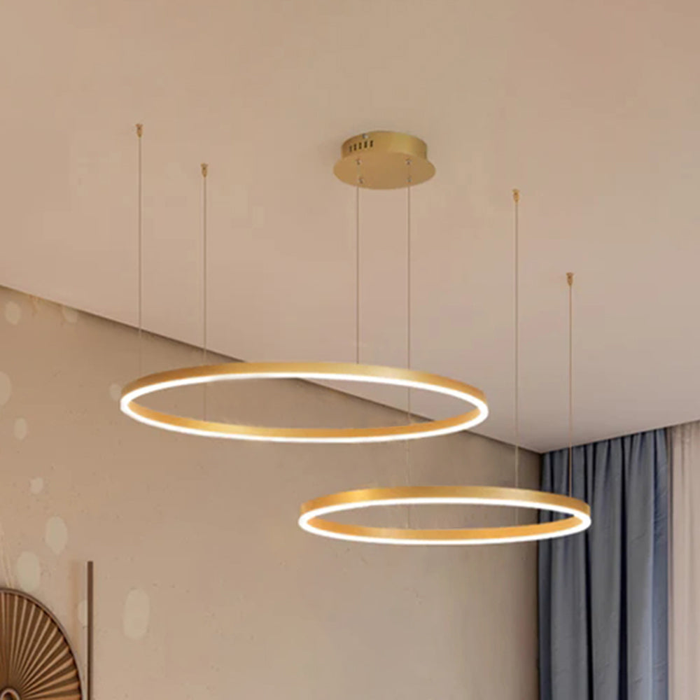 Gold twin-ring pendant light seen from below in a neutral room, showing the staggered ring layout