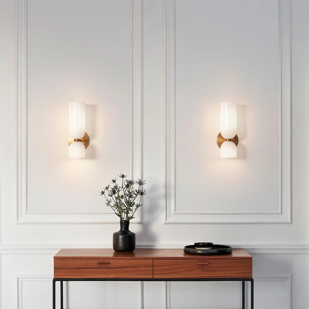 A pair of Solia Twin Glass Wall Sconces lit on a panelled wall above a walnut console table