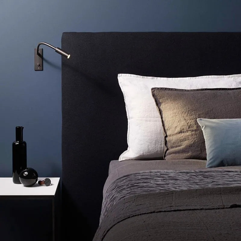 Vexo bronze gooseneck wall light lighting a pillow beside a dark upholstered headboard