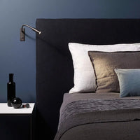 Vexo bronze gooseneck wall light lighting a pillow beside a dark upholstered headboard
