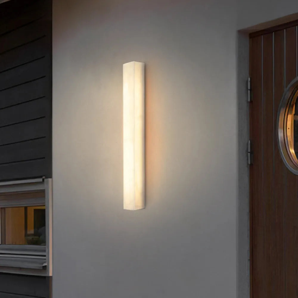 Ayro alabaster wall light lit on a pale exterior wall beside a timber door at night