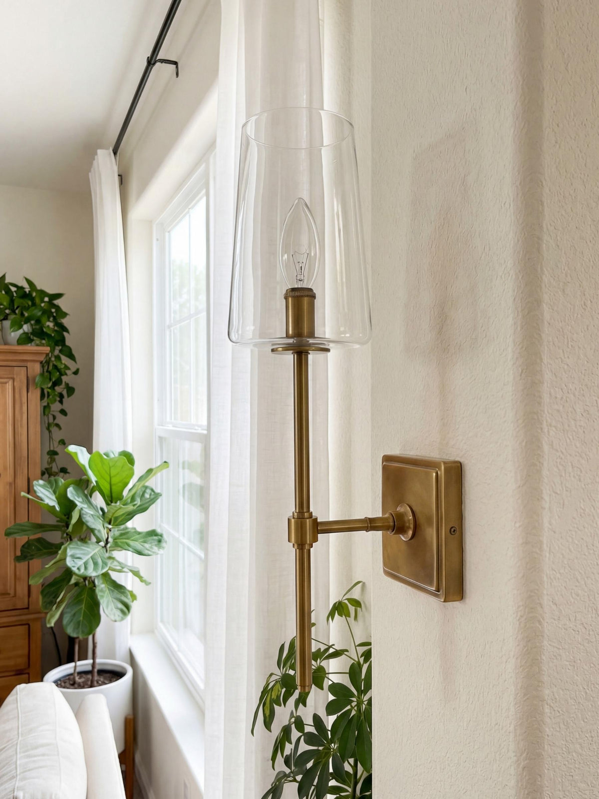 Torin Clear Glass Wall Sconce by a window with sheer curtains, candle bulb unlit