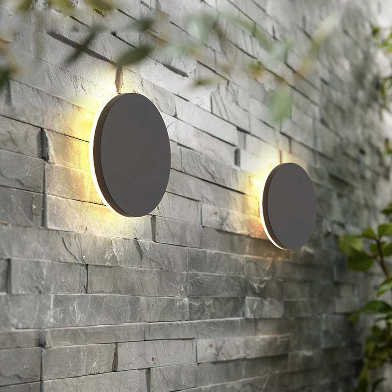 Two Klyno black round outdoor wall lights mounted along a grey stone brick garden wall