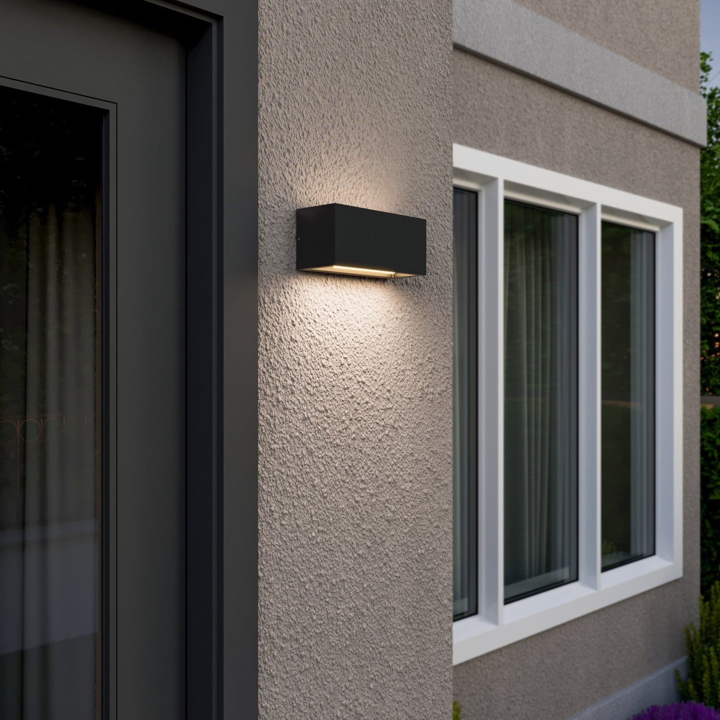 Bexa outdoor wall light switched on at dusk on a house corner beside a window, showing the twin up and down light spread