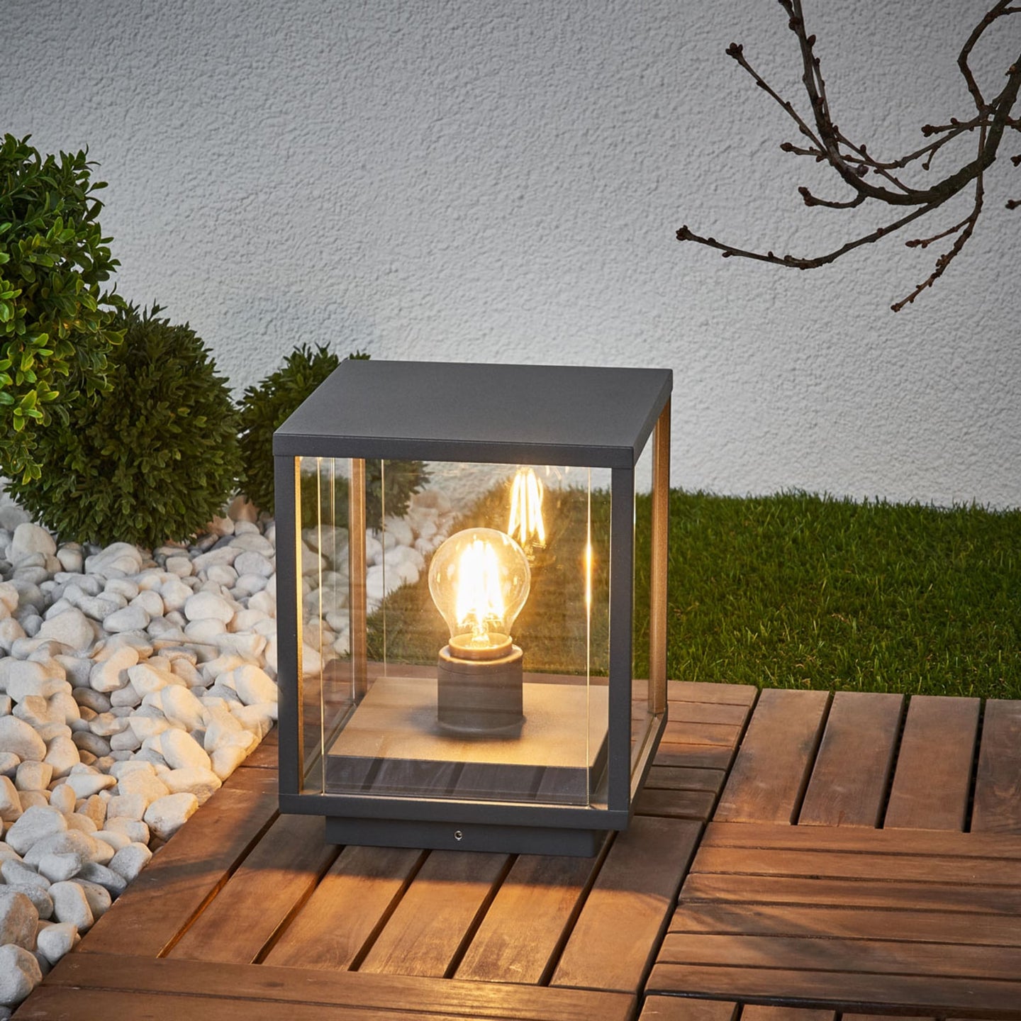 Anle Glass Pillar Light glowing at dusk on decking against a rendered garden wall