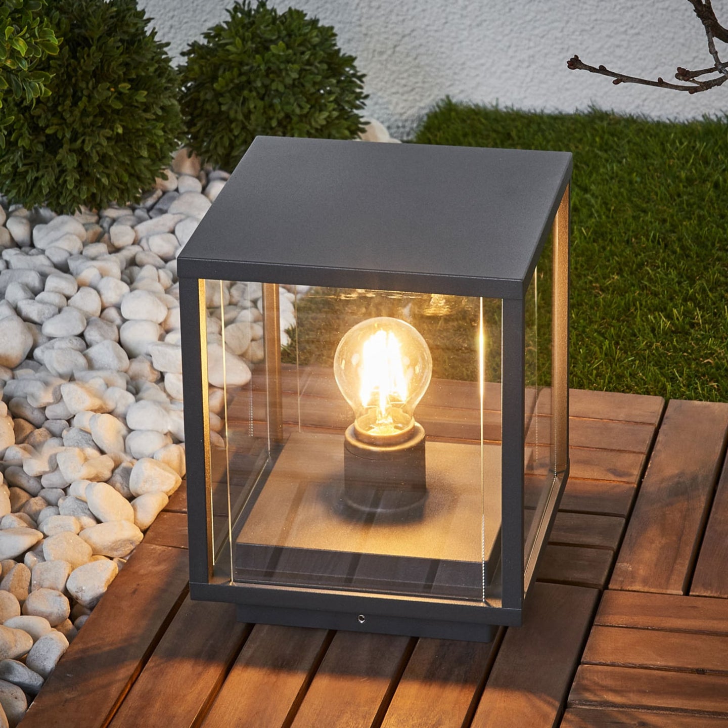 Anle Glass Pillar Light lit on a timber deck beside white pebbles and clipped box