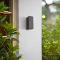 Xato wall light unlit on a pale exterior wall next to garden planting