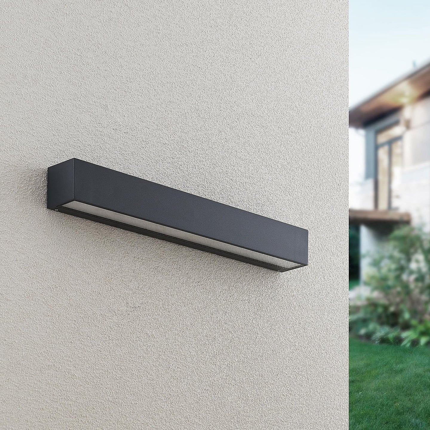 Lino Column outdoor wall light unlit in daylight on a rendered house wall, showing the anthracite finish and slim horizontal profile