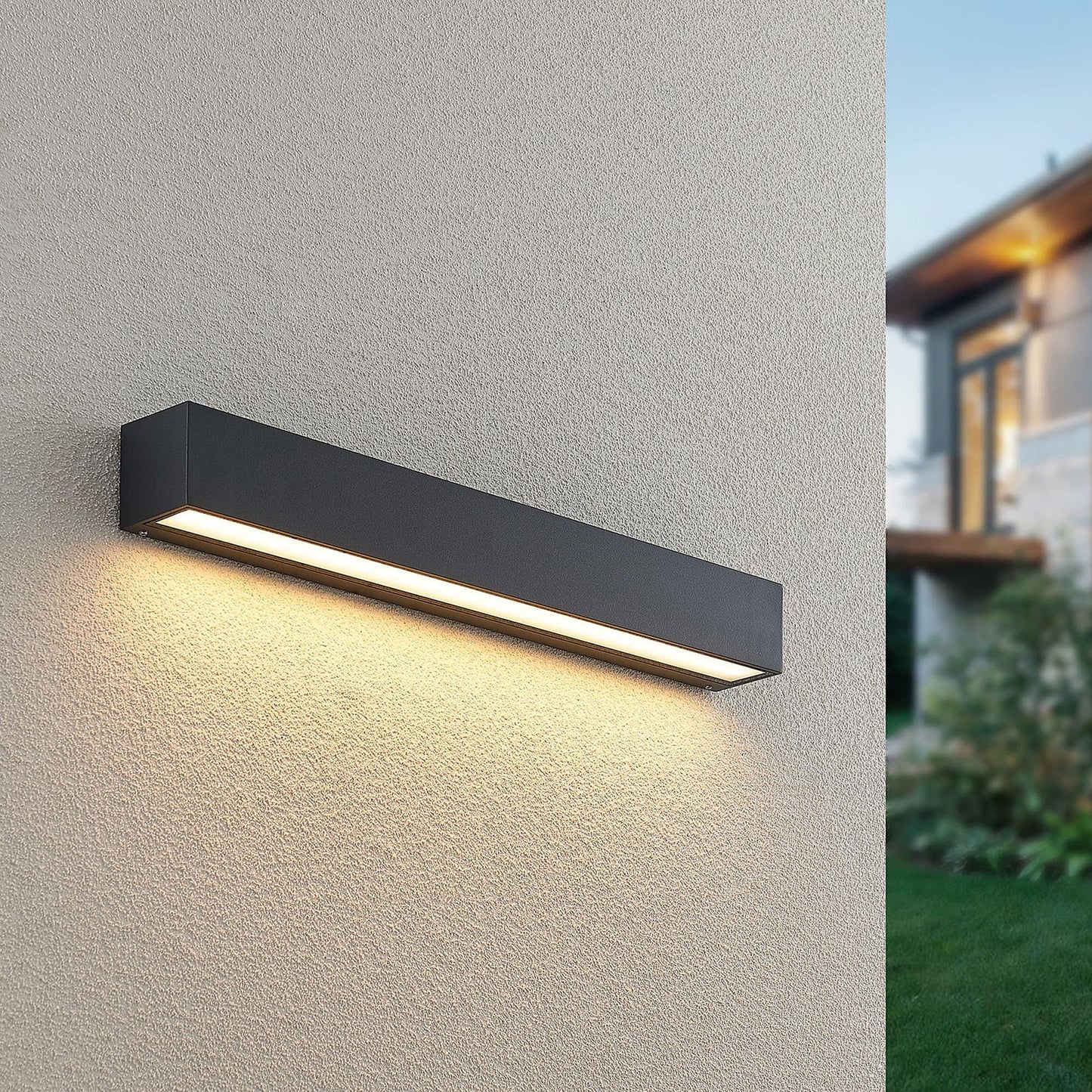 Lino Column outdoor wall light switched on at dusk on a rendered wall, warm light washing down the render beside a garden