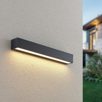 Lino Column outdoor wall light switched on at dusk on a rendered wall, warm light washing down the render beside a garden