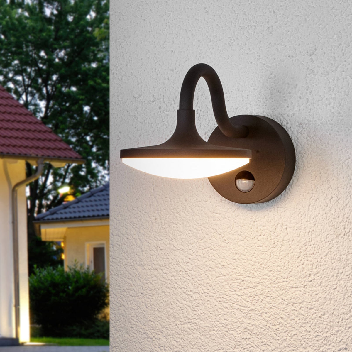 Finno wall light glowing at dusk beside a house with a tiled roof and trees behind