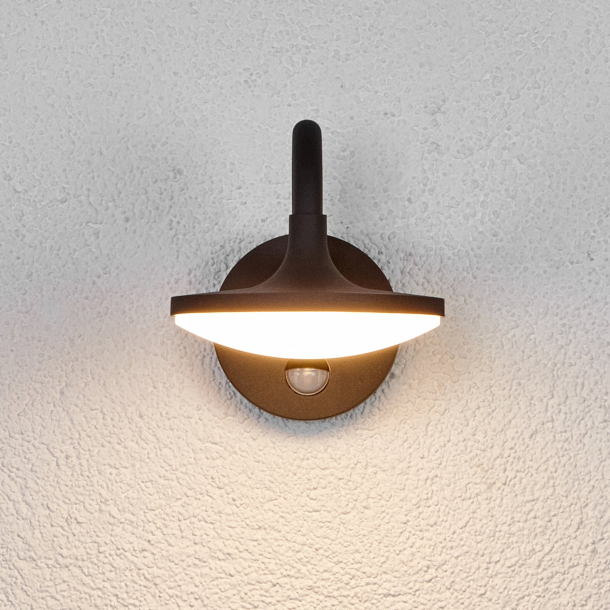 Front view of the Finno wall light lit, showing the disc shade and sensor lens on the mount