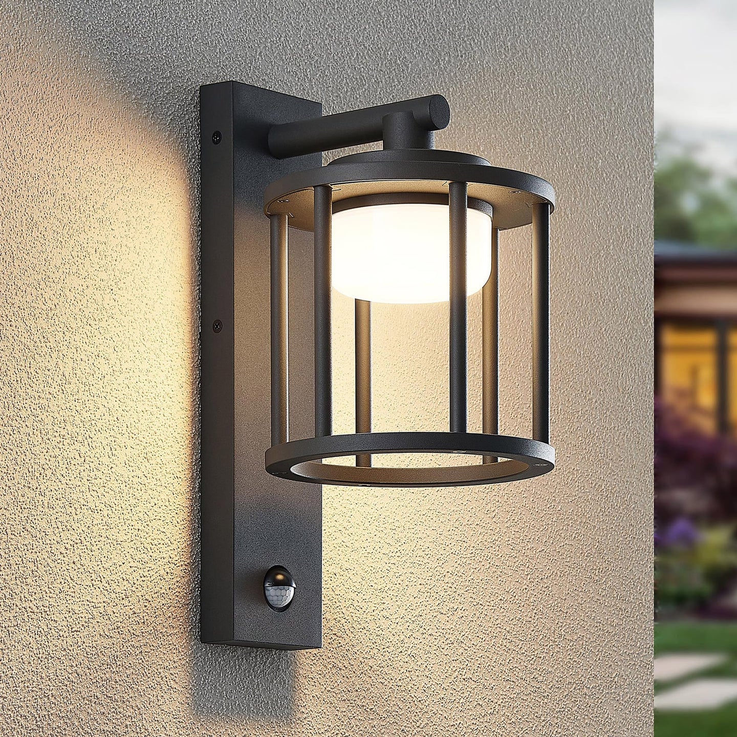 Berno outdoor wall light lit at dusk on a textured house wall beside a garden