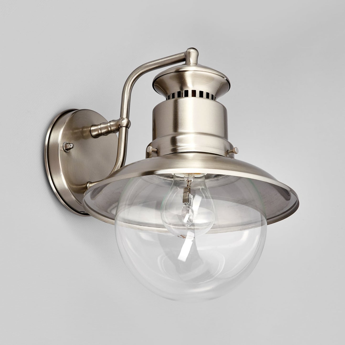 Gwena outdoor wall lantern lit softly against a pale background, showing the clear glass globe and brushed steel shade