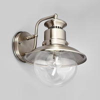 Gwena outdoor wall lantern lit softly against a pale background, showing the clear glass globe and brushed steel shade