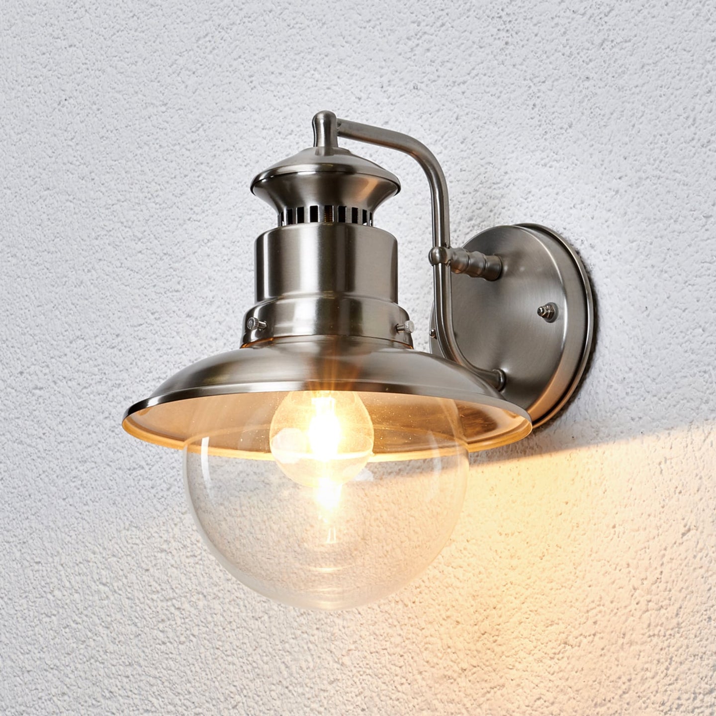 Gwena stainless steel outdoor wall light switched on against a white rendered wall, throwing warm light downwards