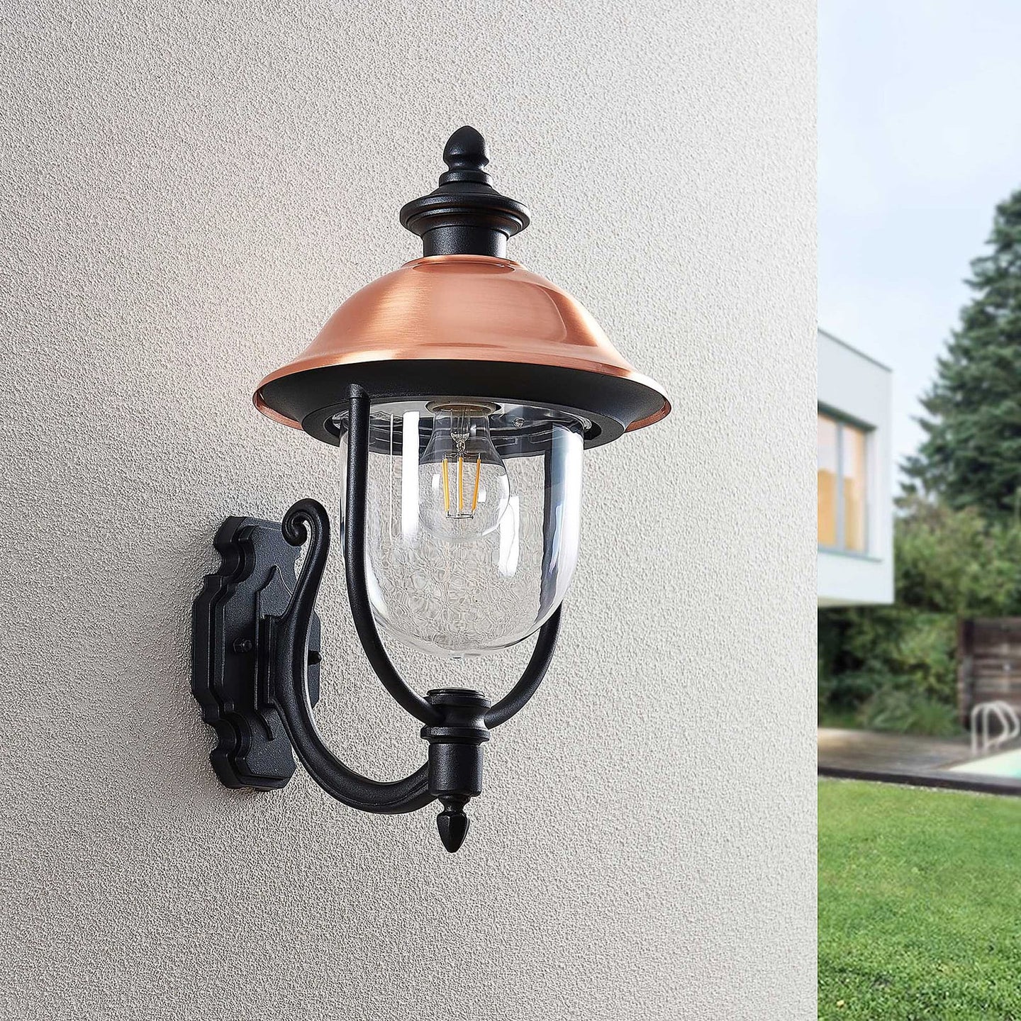 Koma wall lantern shown at an angle on a textured render wall with garden beyond