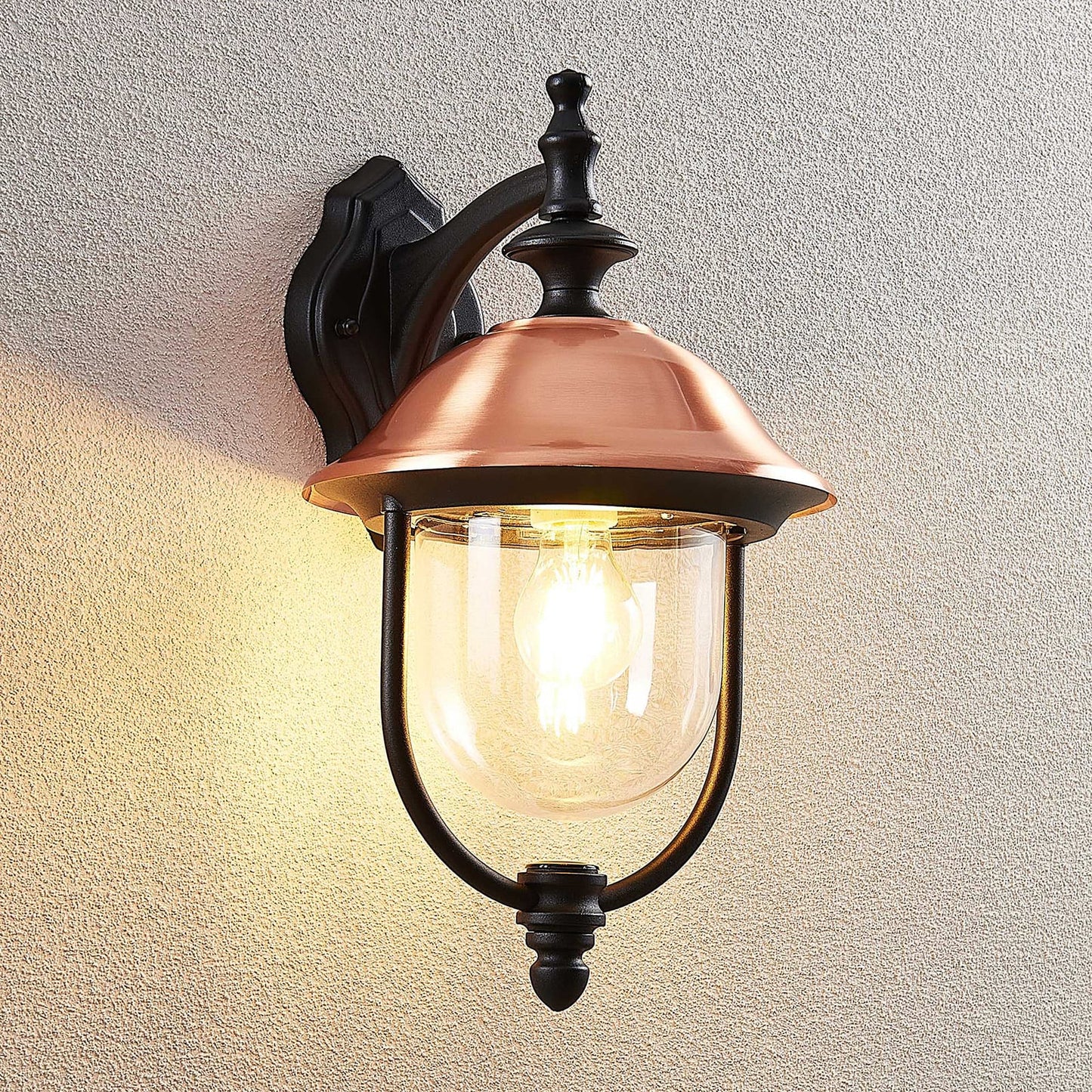 Clivo copper outdoor wall light switched on against a textured rendered wall, casting warm downward light
