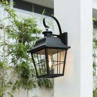 Merto lantern wall light lit with a warm filament bulb beside a white rendered wall and bamboo planting