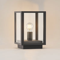 Giva pillar post light lit against a plain pale wall, showing the filament bulb centred on the base plate inside the glass box