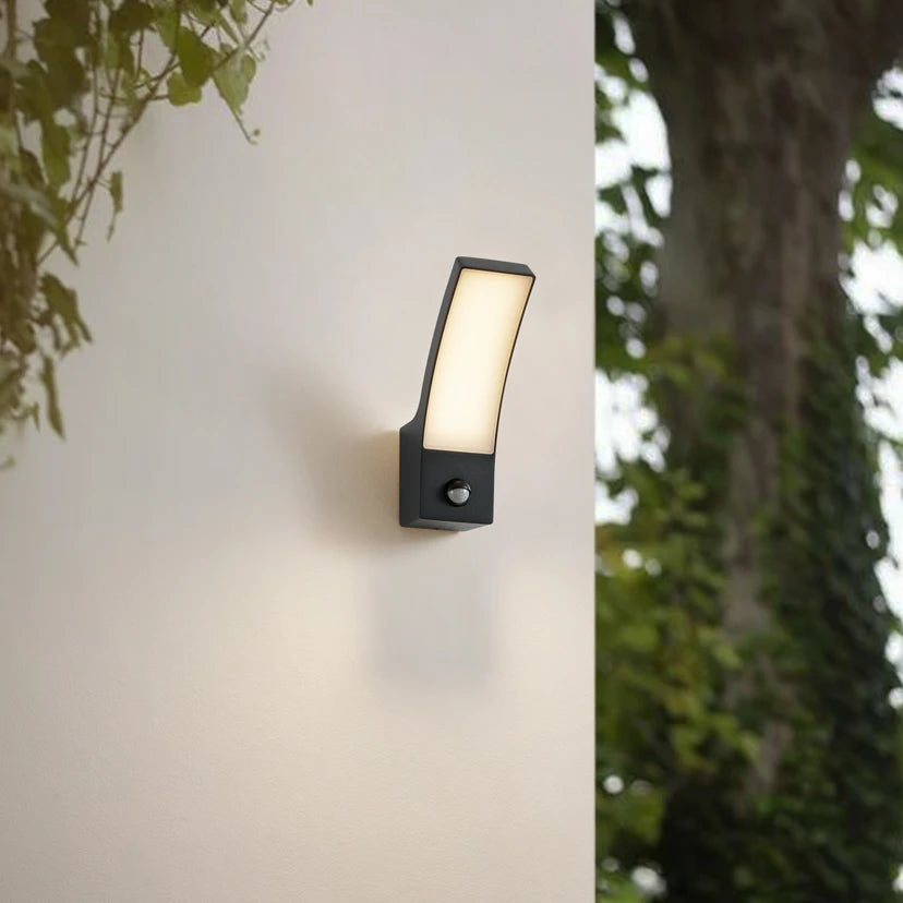 Ilvo sensor wall light glowing on a garden wall with foliage and a tree behind