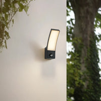Ilvo sensor wall light glowing on a garden wall with foliage and a tree behind