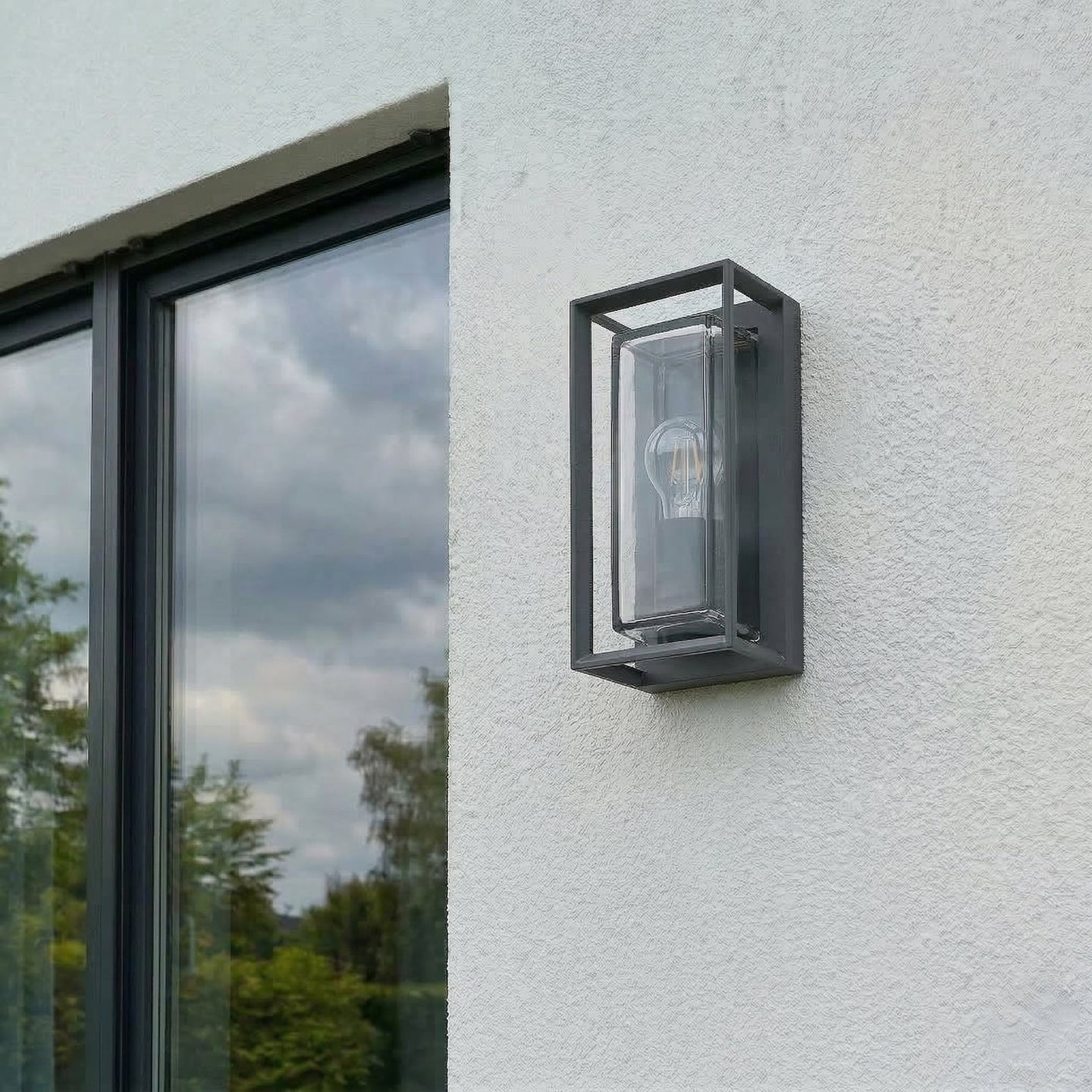 Isra lantern outdoor wall light unlit in daylight on a white house wall, showing the dark grey cage and clear glass against the render