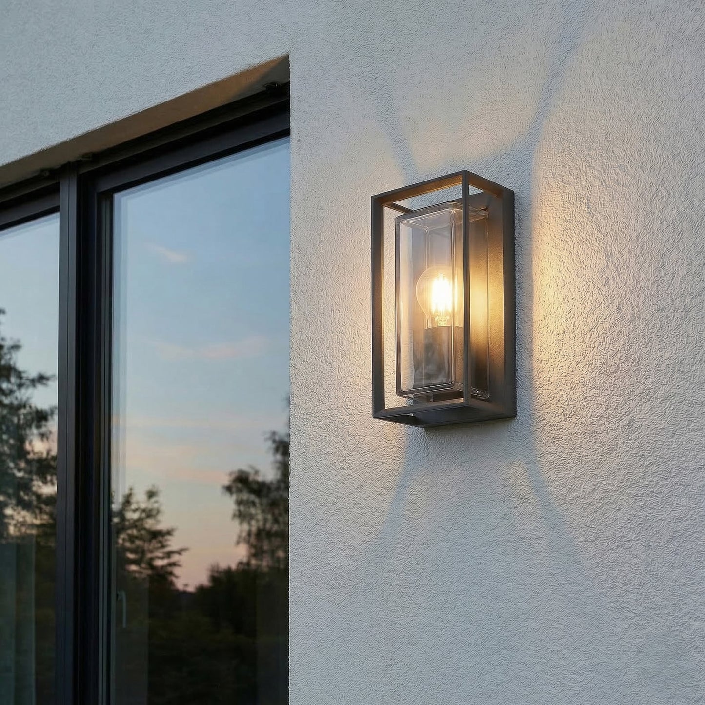 Isra lantern outdoor wall light lit beside a dark framed window at dusk, throwing an even wash across the rendered wall