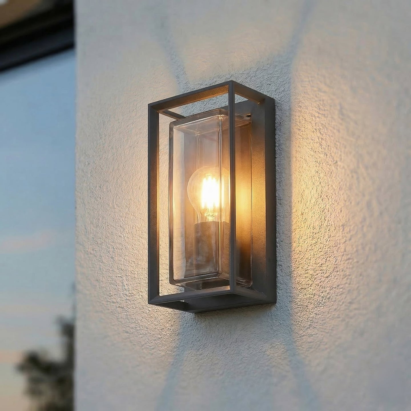 Isra lantern outdoor wall light switched on at dusk on a white render wall, warm light spreading up and down with a soft crossed shadow