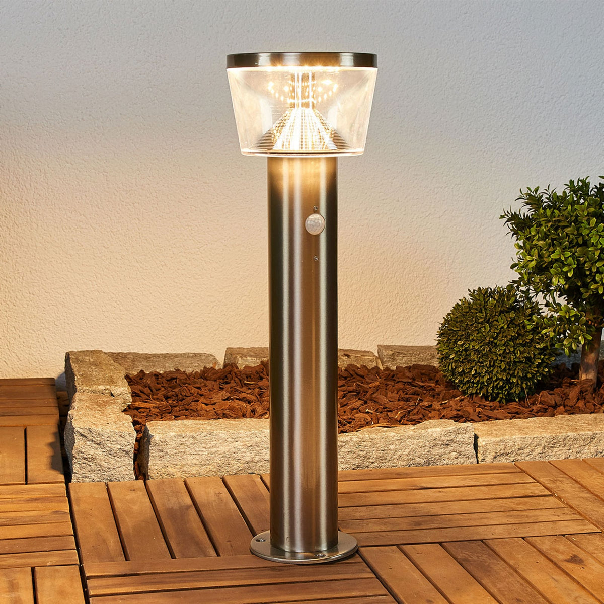 Anjo Solar Pedestal Light lit at dusk beside a garden border