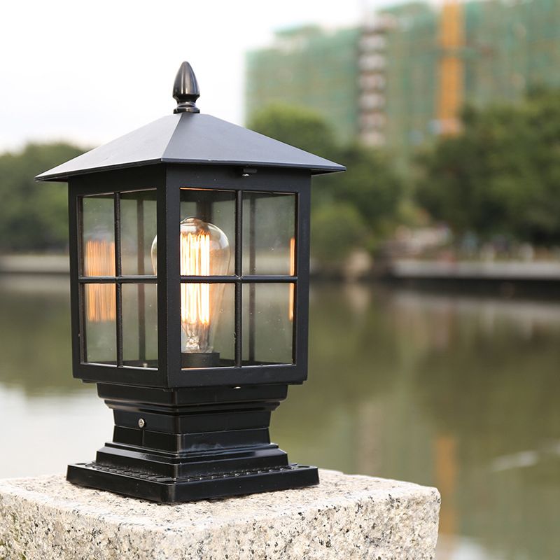 Ashlow black pillar lantern lit on a stone pier beside water, showing the glazed panels and filament bulb