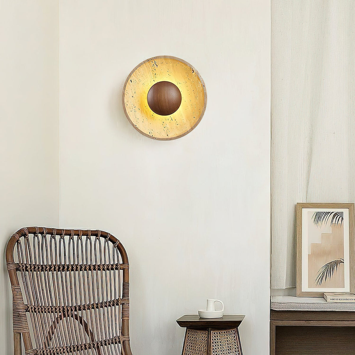 Sunda Travertine Wall Light glowing on a plaster wall beside a rattan chair and framed print