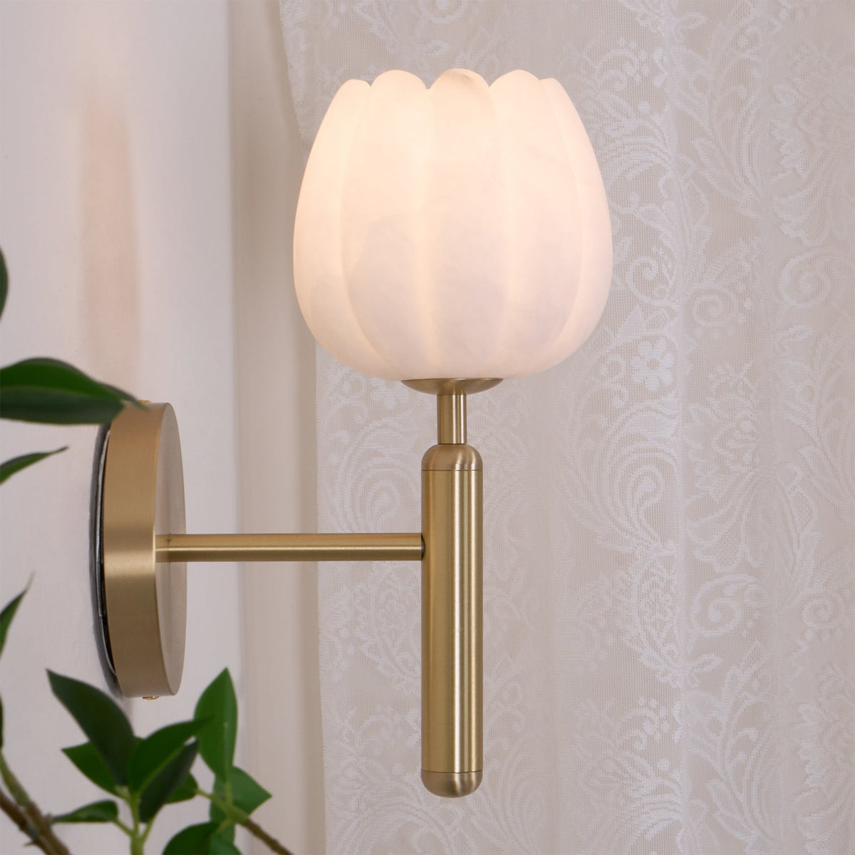 Pellora brass and alabaster wall light lit beside a lace curtain in a bedroom