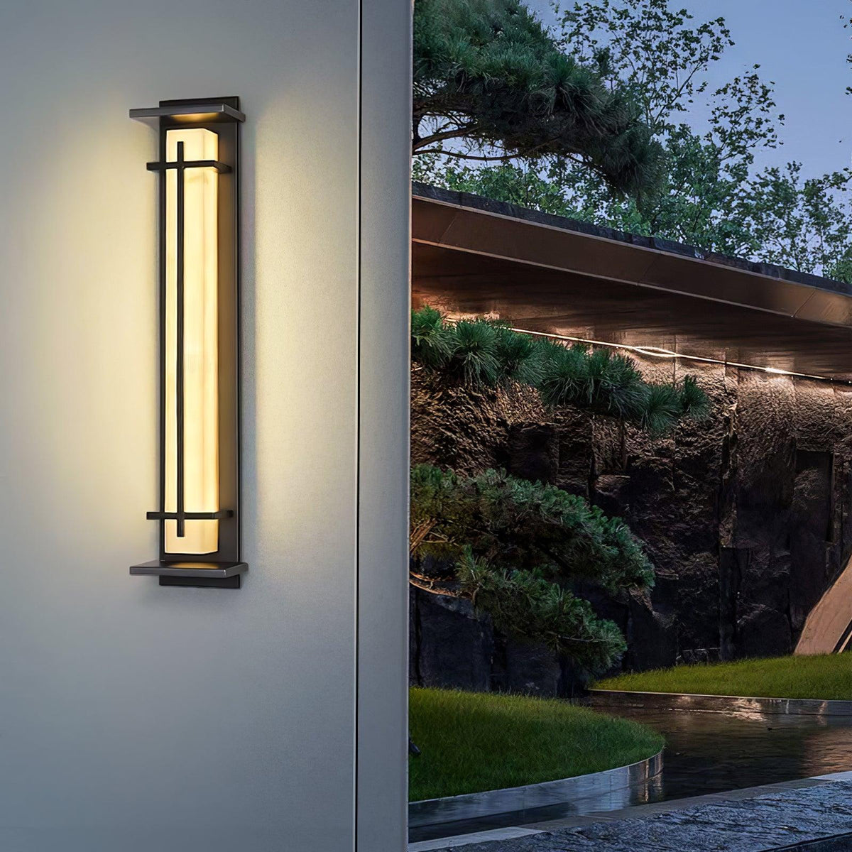 Rayo wall light glowing warm on an exterior wall beside a landscaped rock garden