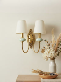 Orla Twin Fabric Shade Wall Light in brass with celadon ceramic accents beside dried flowers on a console