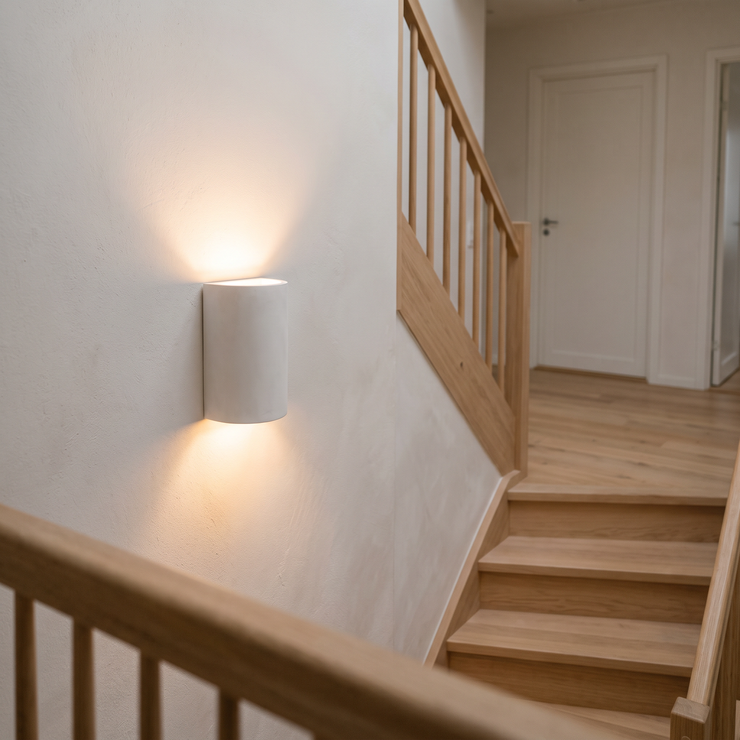 Cova plaster wall light on a staircase wall beside a pale oak handrail, lighting the steps softly