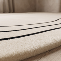 Macro close-up of the Ovala rug surface showing the dense short cream polyester pile and the crisp edges of two fine black border lines