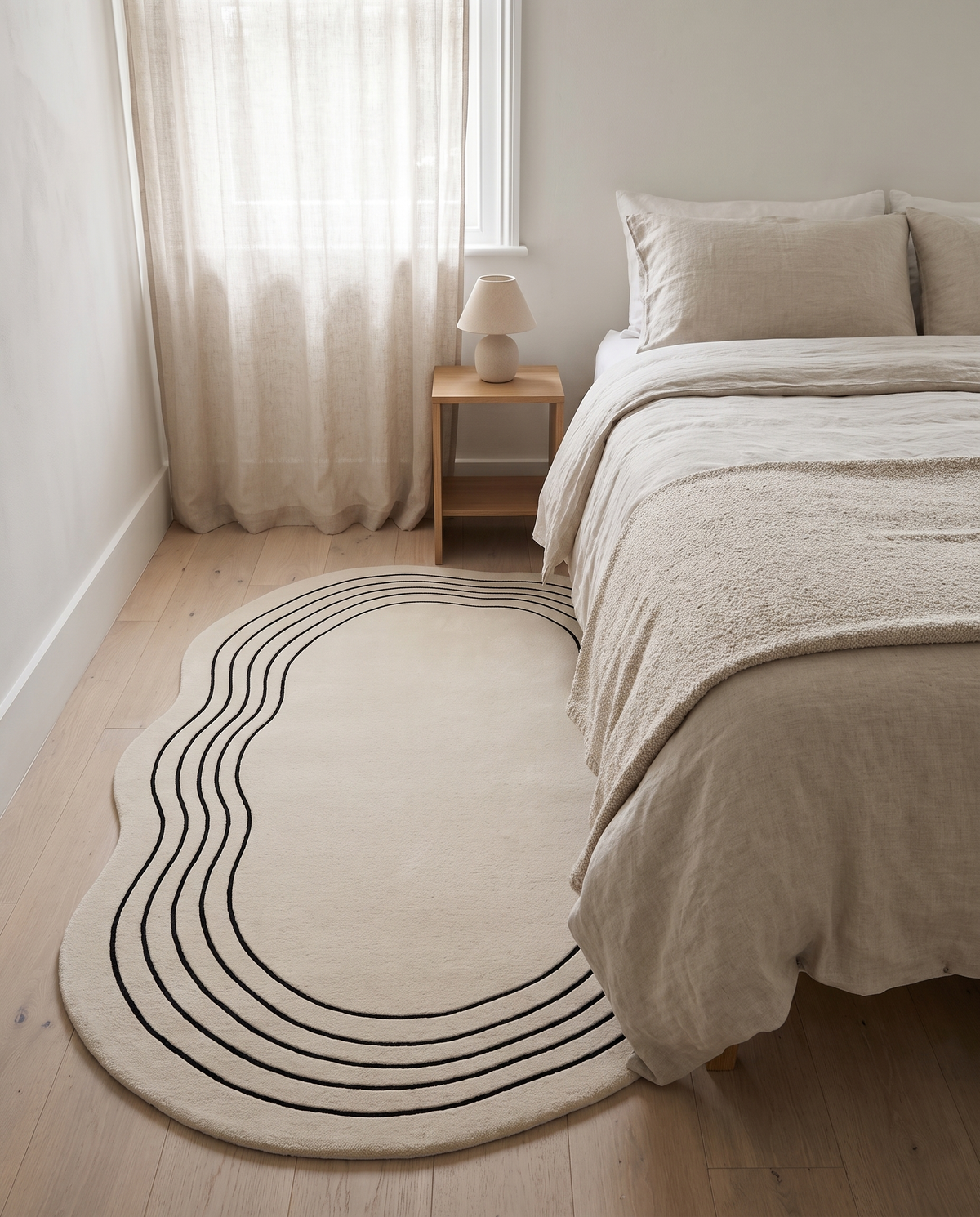 Ovala cream oval rug laid beside a linen-dressed bed on pale oak floorboards in a calm bedroom