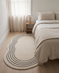 Ovala cream oval rug laid beside a linen-dressed bed on pale oak floorboards in a calm bedroom