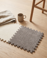 Two Tessa tiles clipped together on a pale oak floor beside a stoneware mug, folded linen throw and oak stool leg showing the real size of a 30 by 30 cm tile