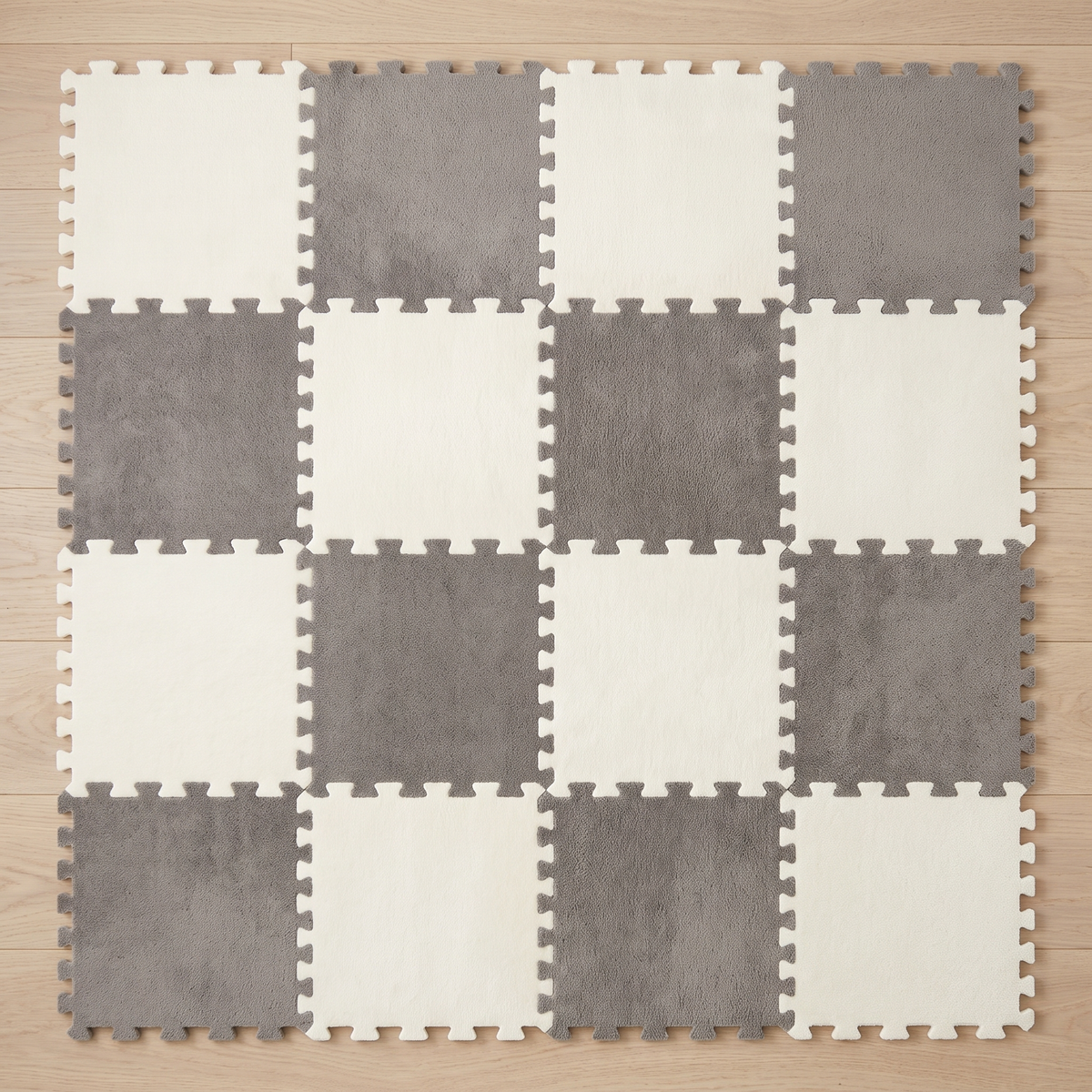 Overhead view of sixteen Tessa modular plush rug tiles assembled into a four by four ivory and grey checkerboard on pale oak floorboards